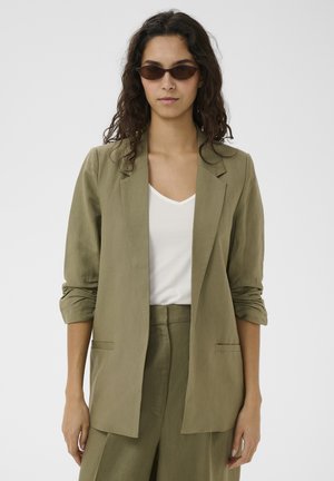 Femeie cu păr lung și creț, purtând ochelari de soare maro, blazer verde măsliniu cu mâneci suflecate, pantaloni asortați și un top alb cu decolteu în V.