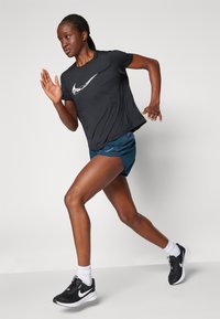 Svart kortermet treningstrøye med hvit logo, blå shorts og svarte joggesko. Modellen løper med en nøytral bakgrunn.