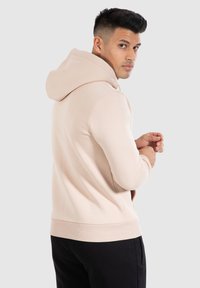 Sweatshirt à capuche rose clair avec une texture lisse, un design ajusté, des poignets et un ourlet côtelés, porté par une personne de dos légèrement tourné.