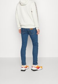 Person med blå slim-fit jeans, en vit hoodie med mönstrade ärmar, och vita-orange Nike-sneakers som står mot en vit bakgrund.