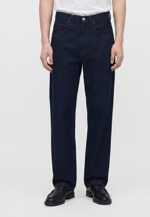 Personne portant un jean droit bleu foncé avec des poches avant, associé à des chaussures habillées noires et une chemise blanche rentrée.