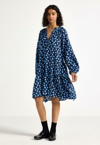 Laivastonsininen mekko sinisellä sydänkuviossa, v-kaula-aukolla, pitkillä hihoilla ja kerroshelman kanssa. Kangas näyttää kevyeltä ja pehmeältä.