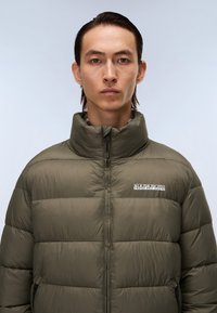 Olive green puffer jacket with a high collar, horizontal stitching, and logo detailing on the left chest; features zippered side pockets.