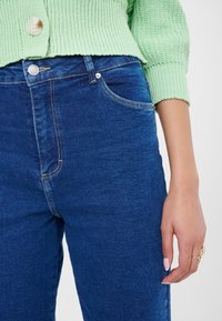 Detailní záběr osoby ve modrých džínách a světle zeleném pleteném svetru s knoflíkem, ruka uvolněně po boku se šperky na prstech.