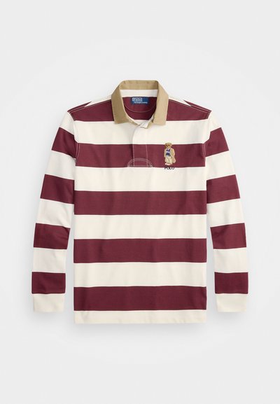 Tricou de rugby cu mâneci lungi, cu dungi orizontale late maronii și albe, guler bej și un urs brodat care poartă un pulover albastru pe piept.