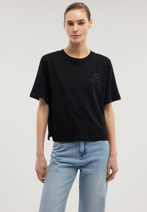 Mujer vistiendo una camiseta negra de manga corta con un pequeño logo y jeans azul claro, de pie contra un fondo liso y claro.