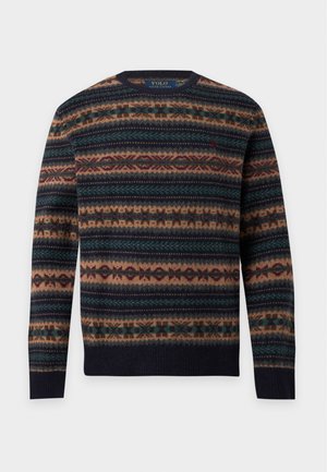 Homme portant un pull sombre à motifs, un pantalon cargo beige avec cordon de serrage et des mocassins beige clair, debout devant un fond blanc uni.
