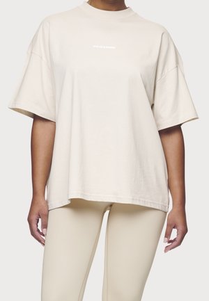 Kobieta ubrana w luźny beżowy T-shirt z krótkimi rękawami oraz dopasowane legginsy na jednolitym tle.