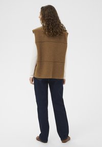 Smeđa bezrukavna prsluk s teksturiranim finišom, s horizontalnim šavovima. Uparen s tamnoplavim ravnim trapericama i smeškim ravnim cipelama.