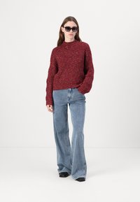 Κόκκινο πλεκτό πουλόβερ με υφή και ψηλό γιακά, συνδυασμένο με ανοιχτό μπλε φαρδύ τζιν. Μαύρα γυαλιά ηλίου ολοκληρώνουν το σύνολο.