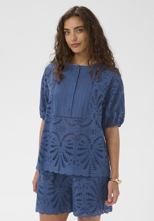 Žena s dlhými kučeravými vlasmi oblečená v modrej blúzke s dierkovanou čipkou s krátkym rukávom a krátkych nohaviciach, pózujúca pred jednoduchým pozadím.