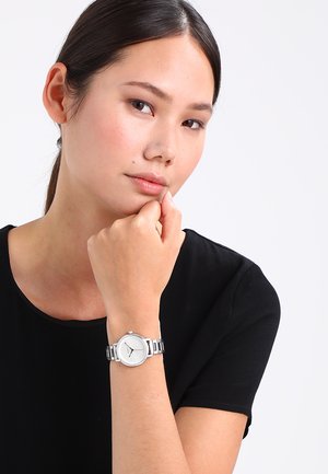 Silberne Analog-Uhr mit rundem weißen Ziffernblatt, Edelstahlarmband, minimalistischer Gestaltung und drei einfachen Stundenmarkierungen; getragen an einem Handgelenk.