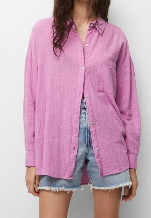 Woman wearing a loose pink textured button-up shirt over light blue frayed denim shorts, standing against a plain background.