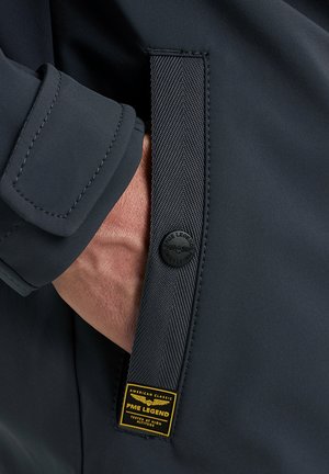 Hand in der Tasche einer dunklen Jacke mit einem strukturierten Riemen, der einen schwarzen Knopf und ein gelbes PME Legend Markenlabel trägt.