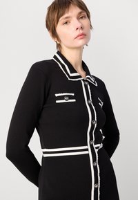 Cardigan noir avec des bordures blanches contrastantes, devant à boutons et deux poches poitrine. Présente un col et une texture côtelée.
