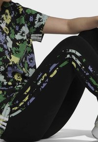 Lyhythihainen vihreä, violetti ja musta kukkakuosiset paita, yhdistetty mustiin leggingseihin, joissa on moniväriset raidat sivuilla.