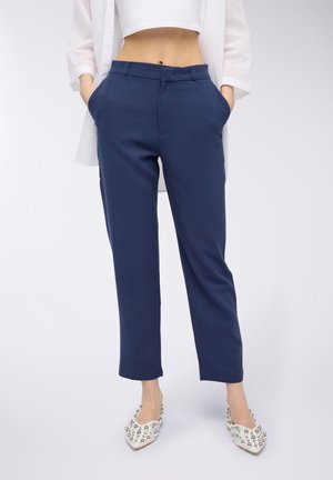Pantaloni blu navy con taglio sartoriale, due tasche frontali e una texture liscia. Indossati con scarpe decorative bianche con accenti metallici.