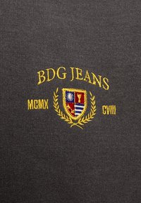 Tessuto grigio con scritta ricamata gialla "BDG JEANS" e un design di scudo con accenti blu, rossi e bianchi, circondato da foglie di alloro.