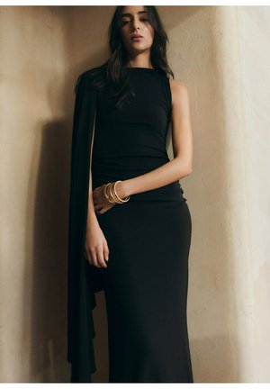 Mujer con un vestido negro sin mangas y una capa drapeada, de pie contra una pared beige, con pulseras doradas en la muñeca.