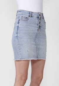 Gonna di jeans azzurro chiaro con una forma aderente, decorata con borchie argentate disposte in un motivo casuale e un orlo sfrangiato. Chiusura a cinque bottoni.