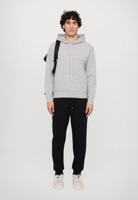 Felpa leggera grigia con cappuccio e lacci, caratterizzata da un logo anteriore, abbinata a pantaloni della tuta neri e sneakers bianche. Abbigliamento atletico semplice e casual.