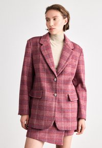 Red plaid blazer with a notched collar, two front pockets, and decorative button detailing; paired with a beige high-neck top.