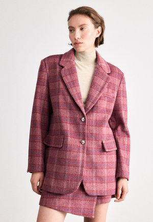Blazer à carreaux rouges avec un col cranté, deux poches avant et des détails de boutons décoratifs ; associé à un haut beige à col montant.