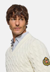 Suéter de punto en color crema con cuello en V, superpuesto a una camisa de rayas en azul claro y blanco; presenta un parche de logo en la manga izquierda.