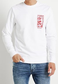 Bílý sweatshirt s dlouhými rukávy a červeným grafickým potiskem na levé straně hrudi. Má kulatý výstřih a texturovanou tkaninu. Nosit s modrými džíny.