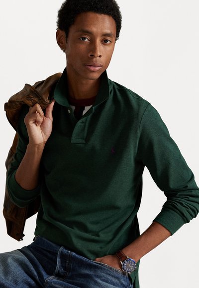 Donkergroene polo met lange mouwen en een kraag, voorzien van een klein paarse logo. Draag het met blauwe denim jeans en een leren jas in het bruin, met een horloge zichtbaar.