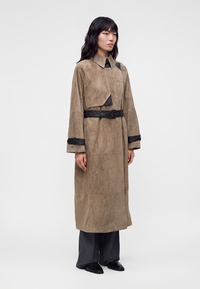 Femme portant un long trench-coat beige en suède avec ceinture noire et détails aux poignets, assorti à un pantalon large foncé et des chaussures noires, debout.