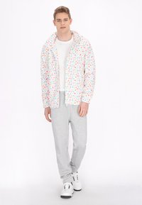 Giacca floreale multicolore con zip frontale, tessuto leggero e cappuccio, abbinata a pantaloni della tuta grigi e sneakers bianche.