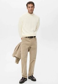 Suéter de punto color crema, pantalones de pana beige, cinturón de cuero negro y mocasines oscuros; el modelo sostiene una chaqueta beige, fondo minimalista.