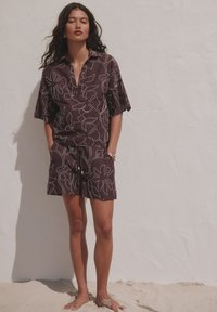 Femme debout pieds nus sur du sable contre un mur blanc, portant une chemise marron à manches courtes à motif floral et un short assorti.