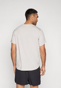 Lys grå t-skjorte med kort erm, laget av teksturert stoff og rund hals, kombinert med mørke shorts. Modell sett fra ryggen.