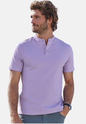 Mann mit lockigem Haar, der ein hellviolettes kurzärmeliges Henley-Shirt und blaue Hose trägt, schaut zur Seite, eine Hand in der Tasche.