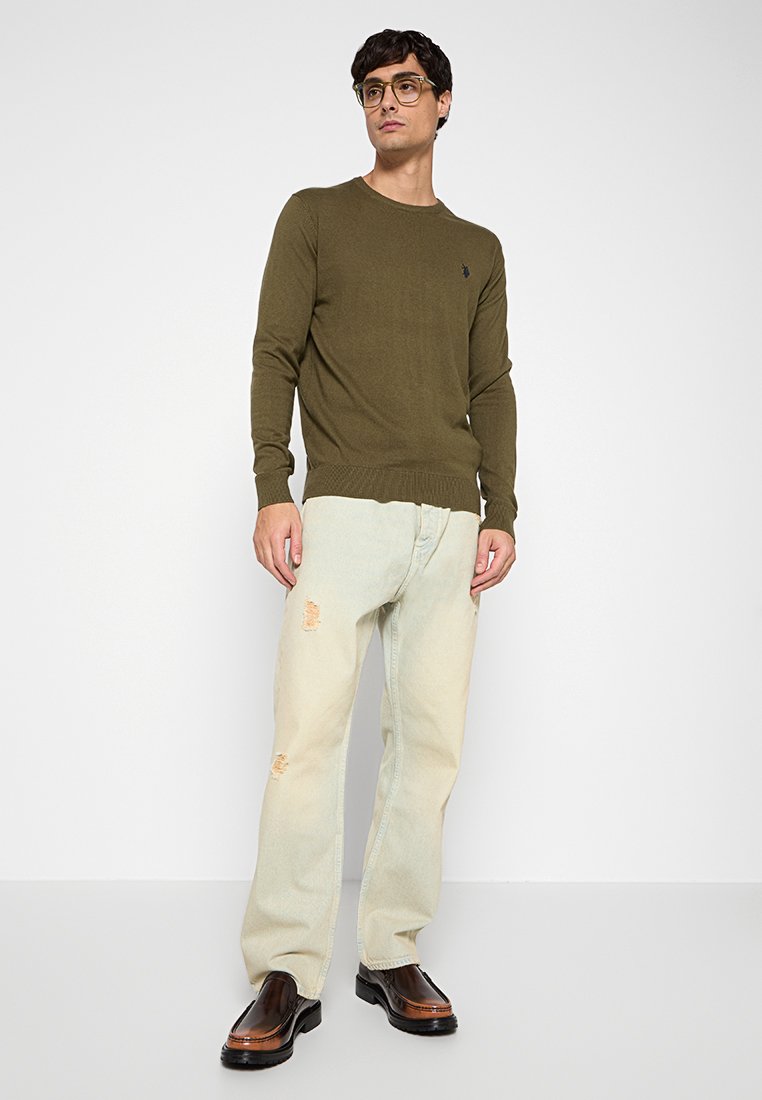 Giovane uomo che indossa un maglione verde oliva, jeans chiari strappati, scarpe di pelle marrone e occhiali, in piedi contro uno sfondo bianco.
