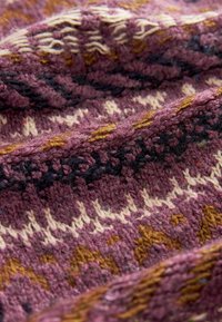 Tissu tricoté en bordeaux profond avec des motifs crème, or et noirs. Fini texturé avec des fibres visibles et des motifs géométriques entrelacés.