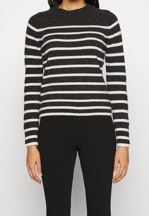 Femme portant un pull à rayures horizontales noires et blanches à manches longues, associé à un pantalon noir côtelé, debout, les bras détendus.