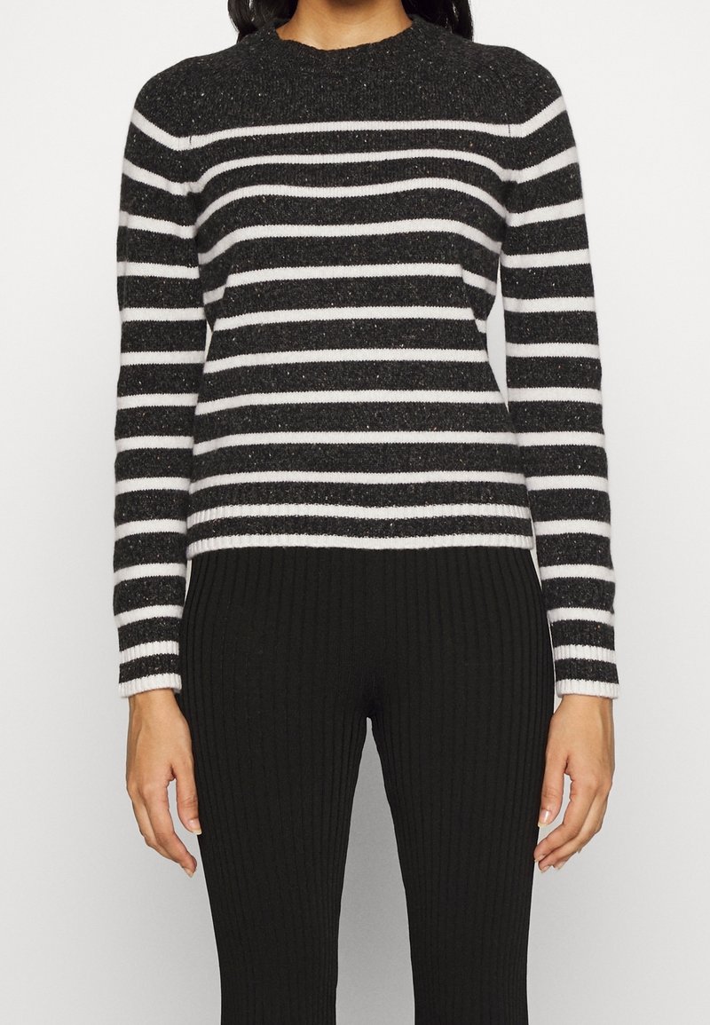 Femme portant un pull à rayures horizontales noires et blanches à manches longues, associé à un pantalon noir côtelé, debout, les bras détendus.