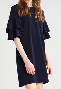 Marineblaue Kleid aus glattem Stoff, mit gestuften Rüschärmeln und einem lockeren Schnitt. Kurze Saumlänge, ohne sichtbare Muster oder Akzente.