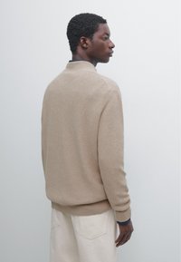 Junger Mann mit kurzen lockigen Haaren steht seitlich, trägt einen beigen Pullover und helle Hosen vor einem schlichten hellen Hintergrund.