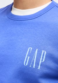 Κοντινή λήψη ενός μπλε t-shirt με στρογγυλή λαιμόκοψη και το λογότυπο "GAP" τυπωμένο σε λευκό στο στήθος, φορεμένο πάνω από ένα λευκό φανελάκι.