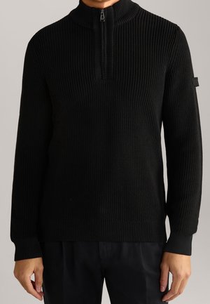 Homme portant un pull en maille côtelée noire avec un col à demi-fermeture éclair et des manches longues, debout devant un fond uni.