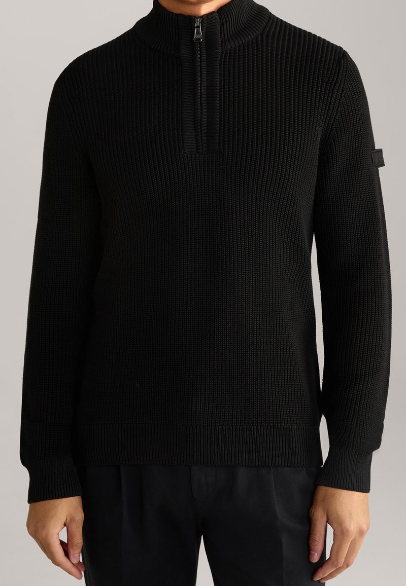 Homme portant un pull en maille côtelée noire avec un col à demi-fermeture éclair et des manches longues, debout devant un fond uni.