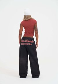Červený těsný crop top v kombinaci se volnými černými džíny s vyšitým červeným logem "Ed Hardy" na zadní straně, se hladkou texturou a širokými nohavicemi.