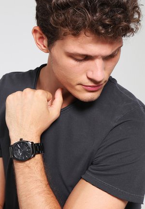 Mann in schwarzem T-Shirt schaut auf die schwarze Armbanduhr an seinem linken Arm, mit lockigem braunem Haar und neutralem Hintergrund.
