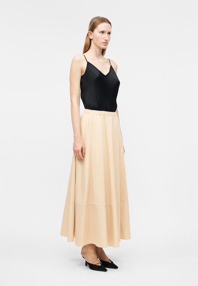 Γυναίκα που στέκεται, φοράει μαύρο αμάνικο τοπ, μπεζ μακρύ φουστάνι σε σχήμα Α και μαύρες μυτερές γόβες σε απλό λευκό φόντο.