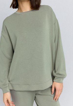 Frau trägt locker sitzendes hellgrünes Sweatshirt und passende Hose, mit schulterlangem dunklem Haar und neutralem Hintergrund.