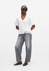 Maglietta bianca con scollo a V e logo "Karl" nero all'orlo, abbinata a jeans in denim grigi sciolti. Gli accessori includono un cappellino bianco e nero e occhiali da sole.